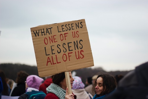Persoon houdt bij een protest een bordje omhoog met daarop de tekst "What lessens one of us, lessens all of us"